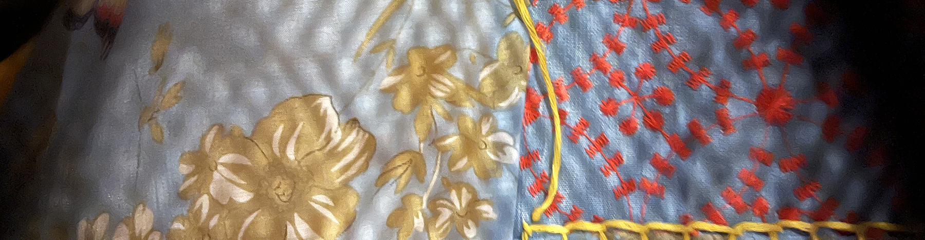 Light blue fabric with gold and red flowers, yellow yarn stitching, and dappled light falling on it with shadows on the right and left edges.